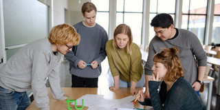 Fünf Studierende stehen um einen Tisch und sehen auf ein Dokument.